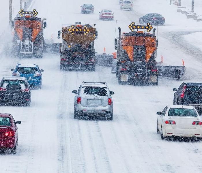 Cars drive behind plows on a highway in a winter storm.